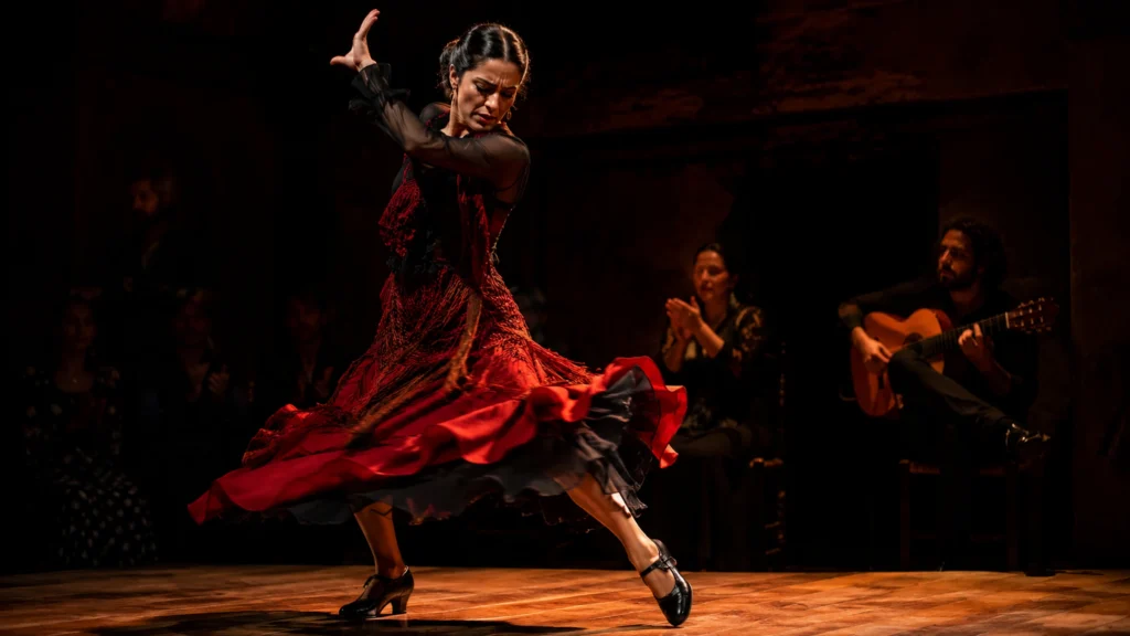 Flamenco tanec na tmavém pódiu, bailaora v červených šatech v dramatickém pohybu a ostrém divadelním světle